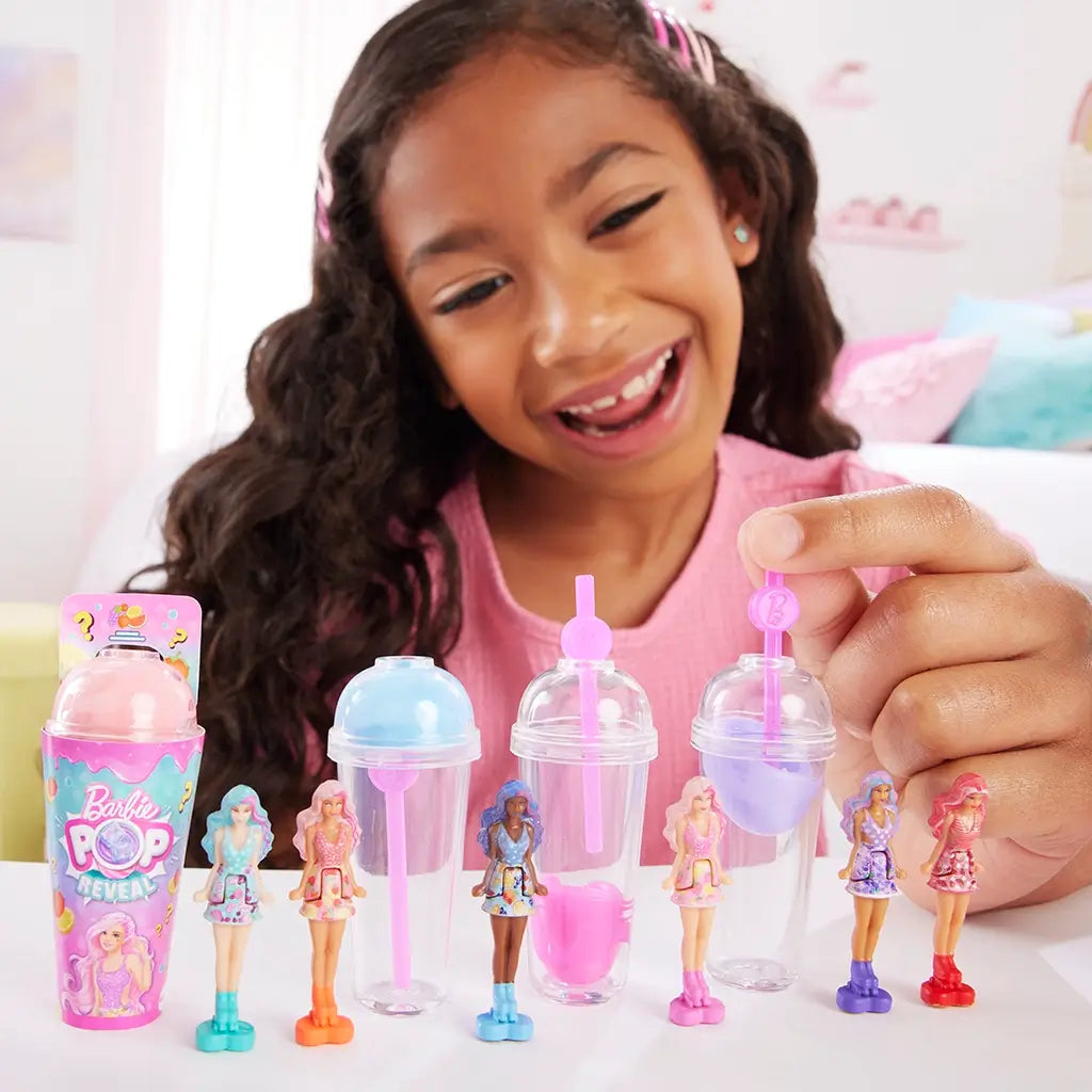 Smiling child plays with Barbie Mini BarbieLand Pop Reveal dolls, unboxing tiny 1.5-inch figures from colorful cup containers and arranging them in a row on a table.