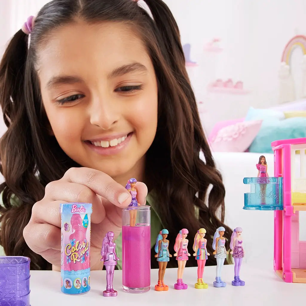 Smiling child reveals Barbie Color Reveal mini dolls in pink water tube, displaying multiple colorful mini fashion dolls lined up alongside a dollhouse for creative storytelling play.