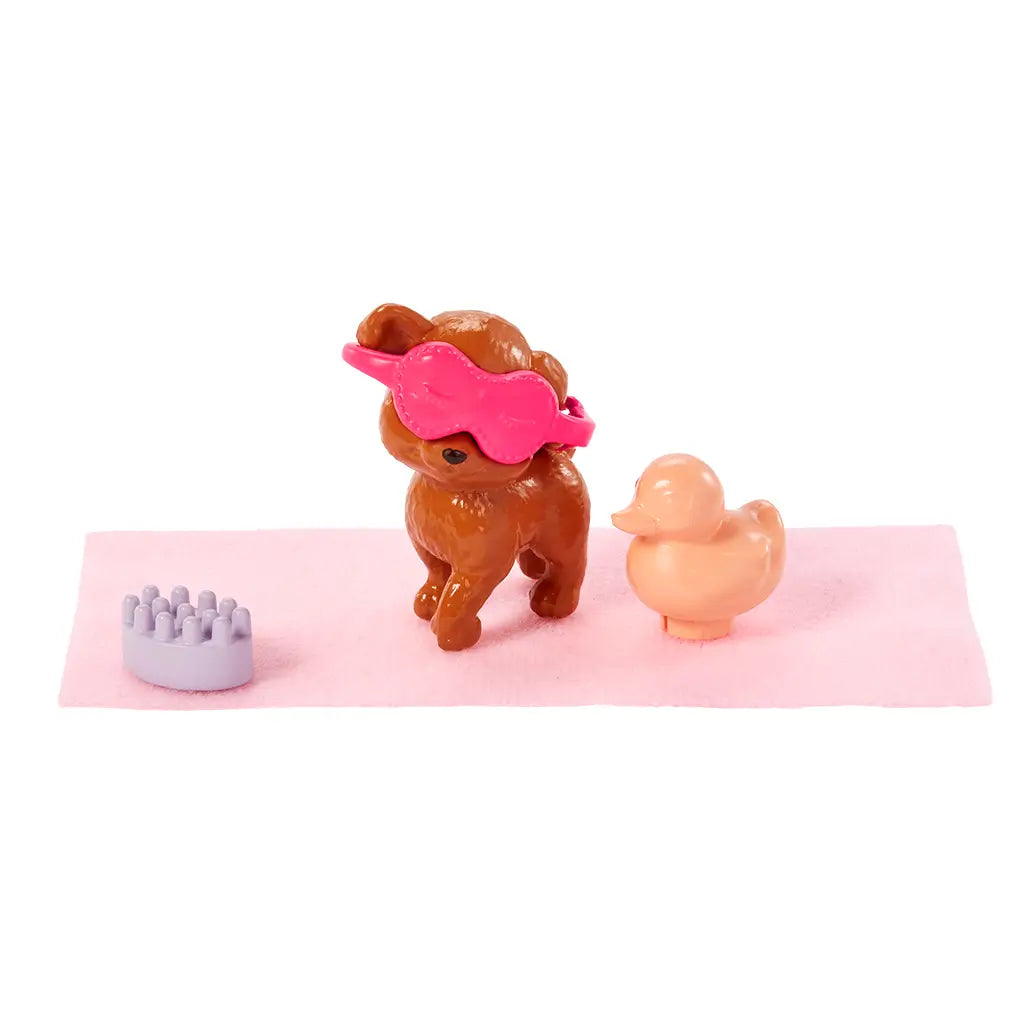 Small brown puppy toy with a pink sleep mask, peach rubber duck, and lilac massager placed on a soft pink towel, part of Barbie’s spa day playset.