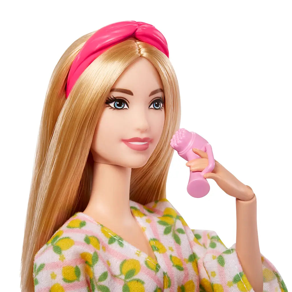 Close-up of Barbie doll wearing a lemon-print robe and pink headband, smiling while holding a pink facial massager accessory near her cheek.