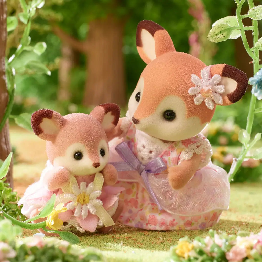 Calico Critters Deer Baby and mother figures posed in close-up, both in floral pastel dresses with lace details, standing in a forest scene surrounded by greenery and small blooming flowers.