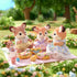 Calico Critters Deer Family toy figurines enjoying a picnic outdoors on a pink blanket with miniature food, surrounded by green grass, trees, and colorful flowers.