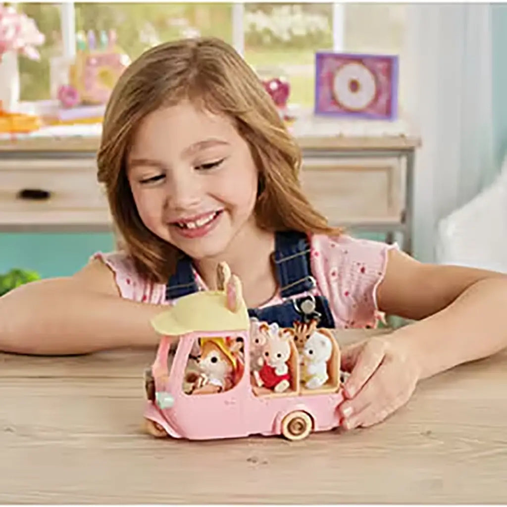Smiling child in a pink shirt and denim overalls plays with the Calico Critters Dip Dip Donuts wagon, a pastel pink truck carrying small animal figures in the back seats on a wooden table.