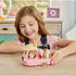 Smiling child in a pink shirt and denim overalls plays with the Calico Critters Dip Dip Donuts wagon, a pastel pink truck carrying small animal figures in the back seats on a wooden table.
