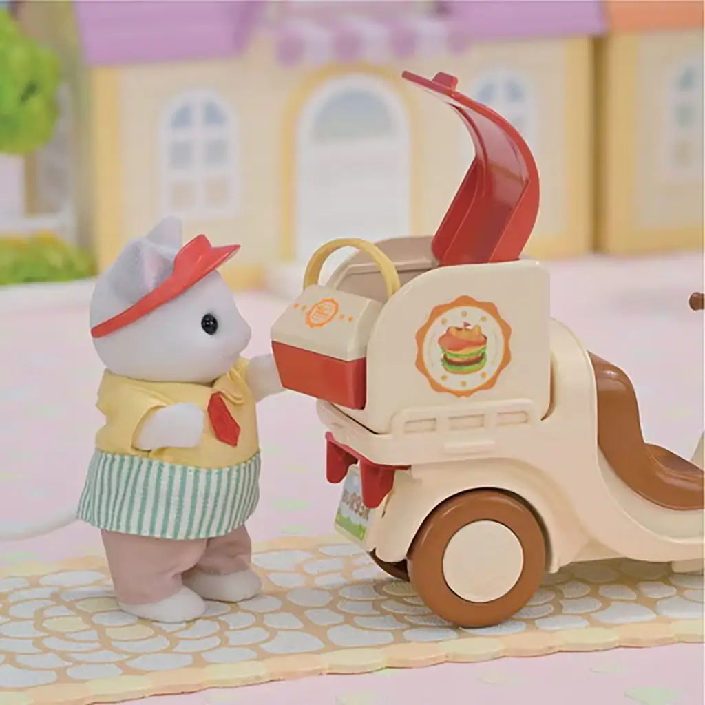 Close-up of Calico Critters cat figure in uniform placing food into a cream and brown scooter delivery box with a burger logo, set in a pastel village background.