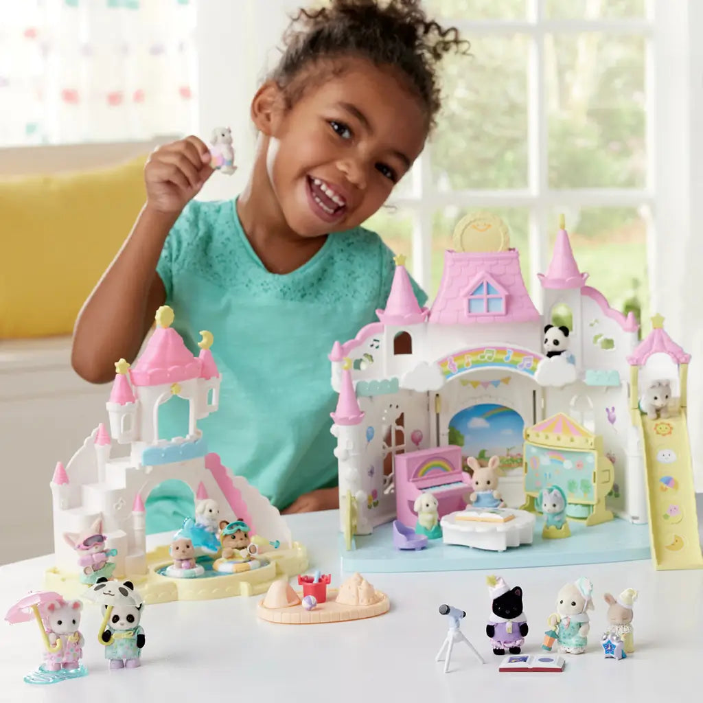 Smiling child in a teal shirt plays with Calico Critters castle playset featuring pastel towers, animal figures, water play area, and accessories including the Pool Fun Trio.