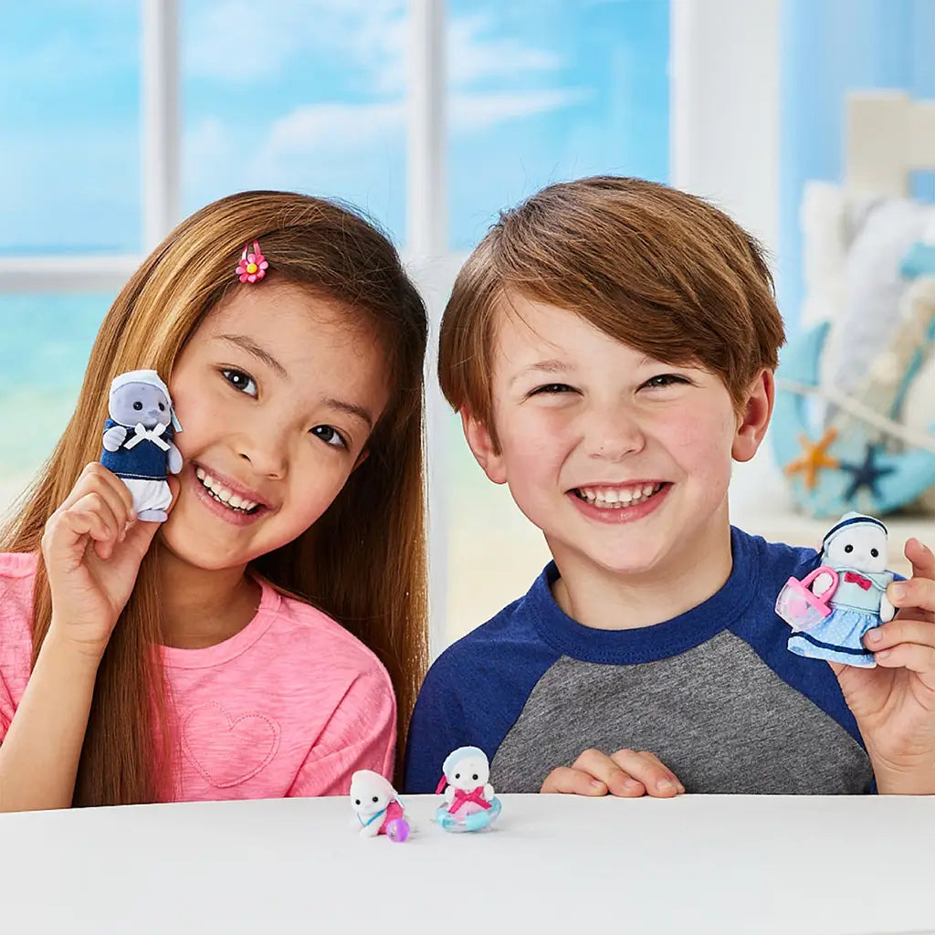 Calico Critters Seal Family toys shown with a smiling boy and girl, each holding plush seal figures dressed in blue and pink outfits, posed indoors with bright window light.