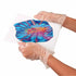 Gloved hands tilt a small white canvas covered in bright blue, red, and yellow acrylic paint swirls, showing the paint-pouring effect from the Palmer Paints Pouring Art Kit.