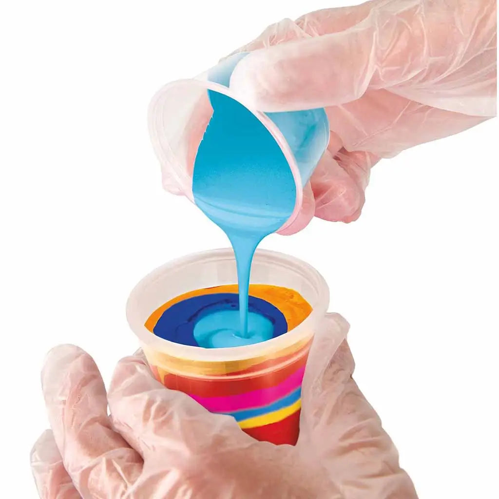 Hands wearing plastic gloves pour turquoise acrylic paint into a clear cup layered with orange, yellow, and pink paints, demonstrating the color layering method in paint pouring.