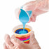 Hands wearing plastic gloves pour turquoise acrylic paint into a clear cup layered with orange, yellow, and pink paints, demonstrating the color layering method in paint pouring.