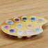 Yellow plastic palette holding multiple small acrylic paint pots in pastel and bold colors with numbered lids, neatly arranged on a wooden table for organized painting sessions.