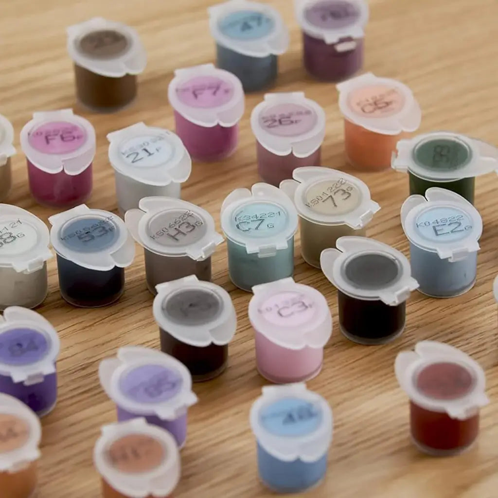 Assorted acrylic paint pots in vibrant shades including pink, blue, green, and gray, each with numbered lids, scattered across a wooden surface for creative paint by numbers projects.