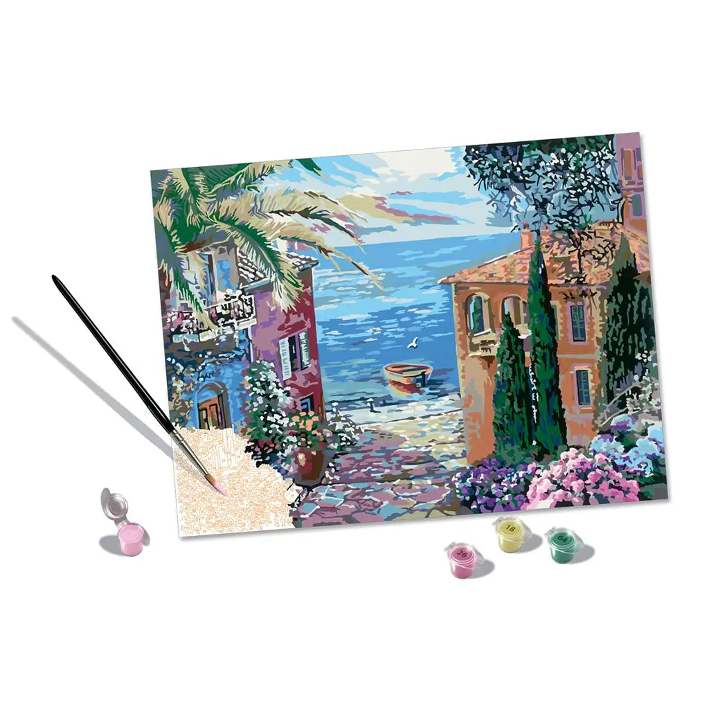 Mediterranean landscape paint by numbers set displayed with finished canvas, included acrylic paint pots, and a double-ended paintbrush arranged against a white background for product showcase.