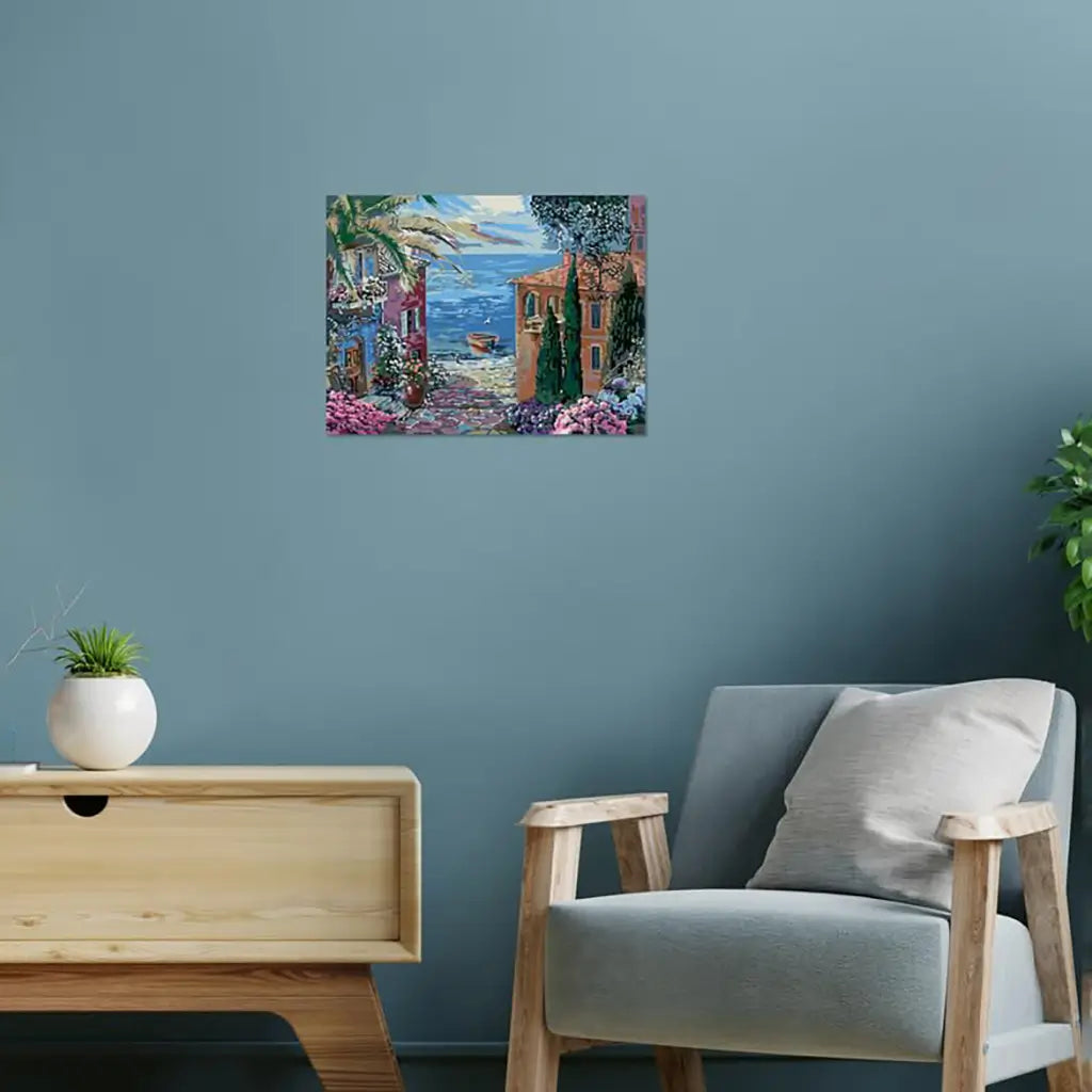 Framed Mediterranean landscape paint by numbers artwork hung on a light blue wall above a wooden cabinet and beside a cushioned chair, styled as home décor in a modern living space.
