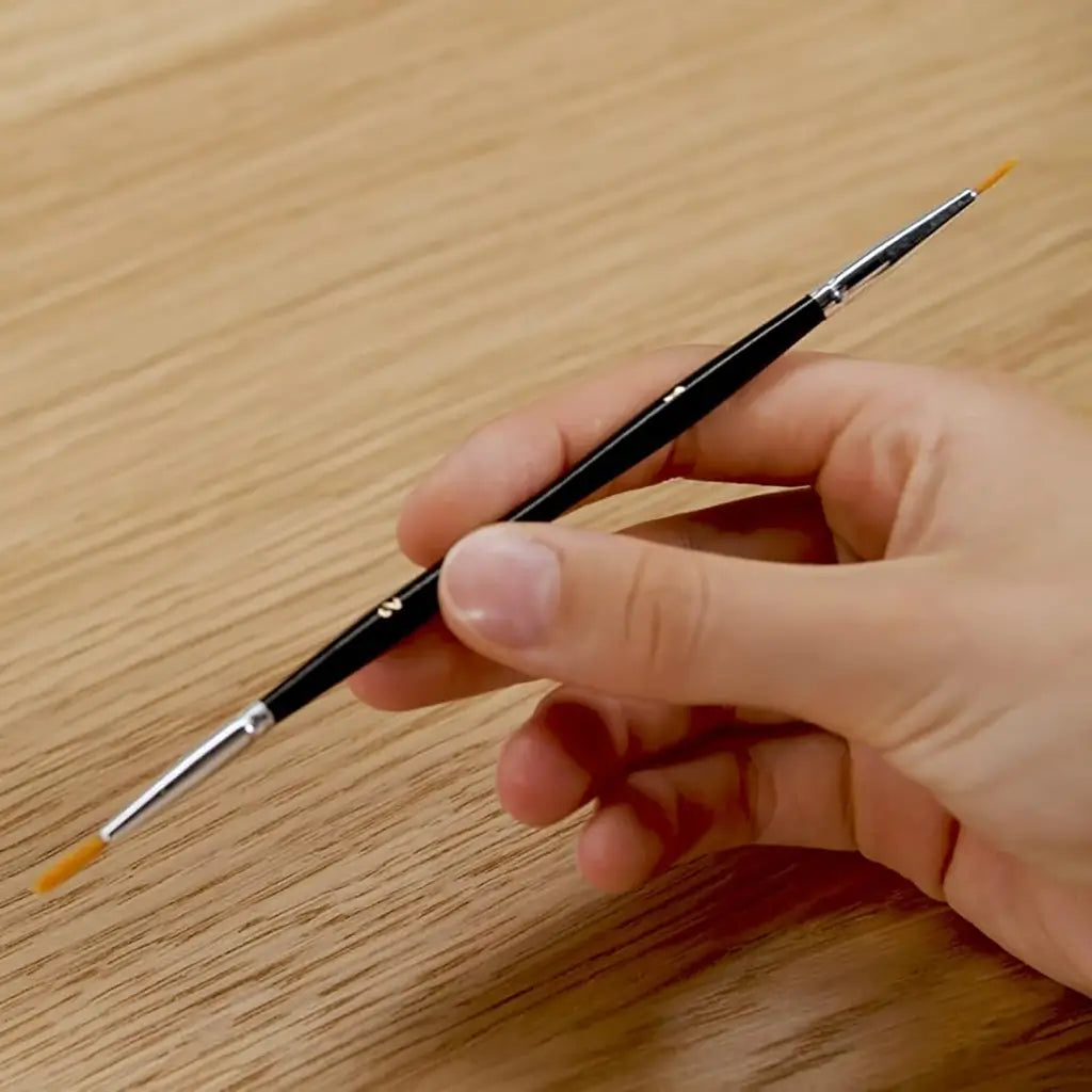 Black double-tipped paintbrush with fine orange bristles held in hand, designed for precise acrylic painting on wood or canvas surfaces, ideal for paint by numbers kits.