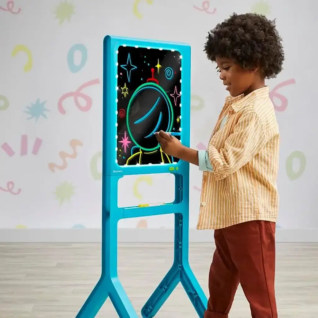 Young child drawing on the Discovery Glow Easel standing art board, creating a neon astronaut design on the illuminated surface in a playroom with colorful wall patterns.