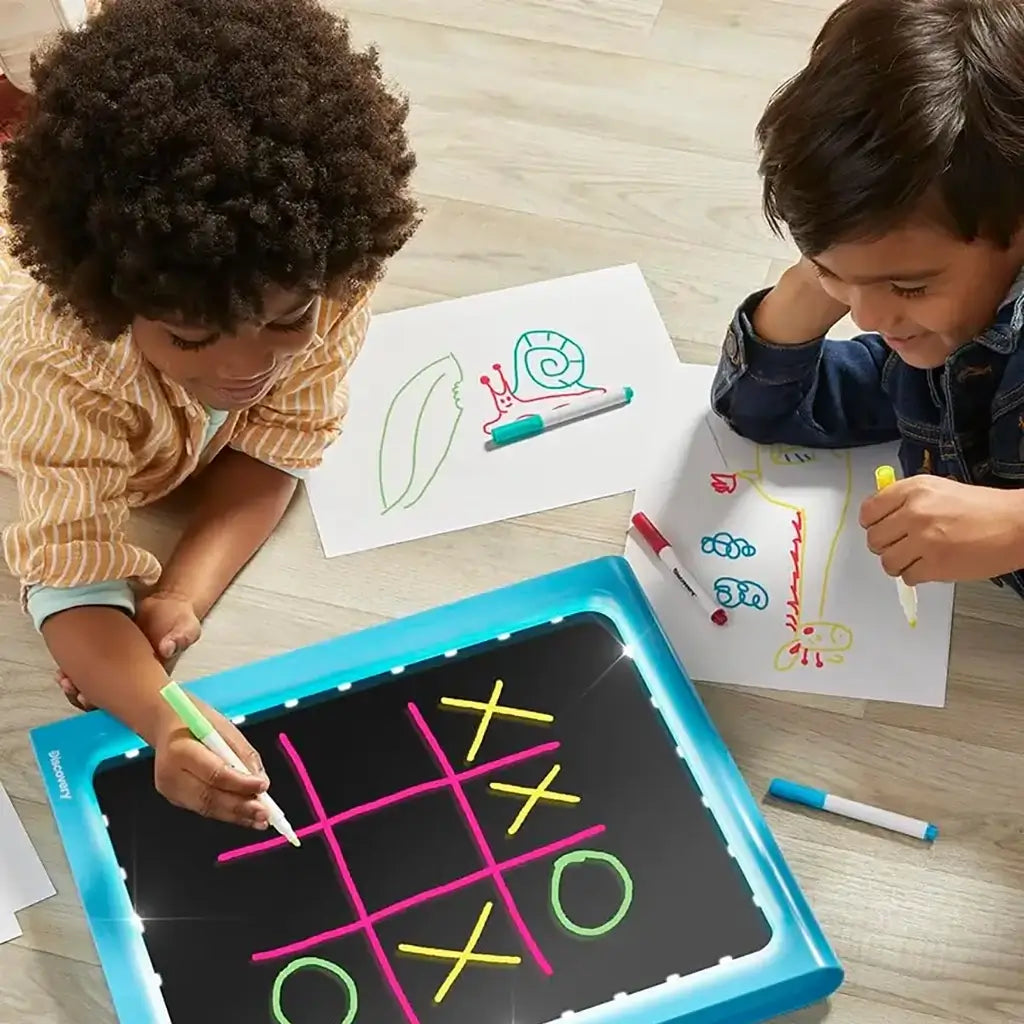 Two children drawing with Discovery Glow Easel light board and markers, one creating a tic-tac-toe game while the other sketches colorful animals on paper beside it.