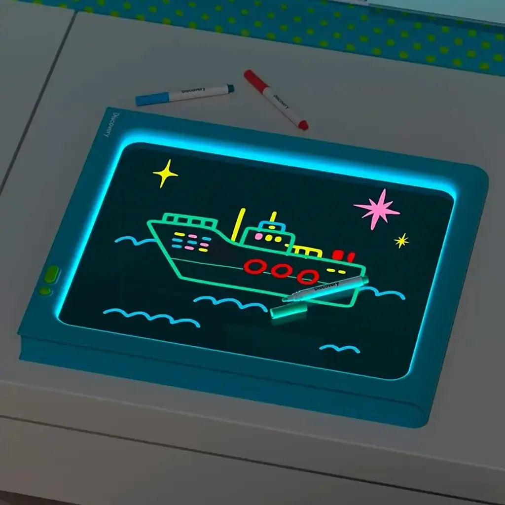 Discovery Glow Easel LED light drawing board lying flat on a desk, glowing neon artwork of a ship and stars, with blue and red markers placed nearby under dim lighting.