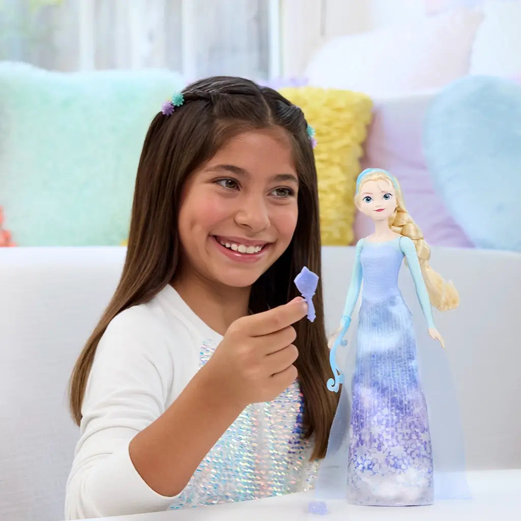 Smiling girl playing with Disney Frozen Elsa doll in shimmering blue gown, holding icy accessory in a cozy playroom setting with pastel pillows, creating magical Frozen moments.