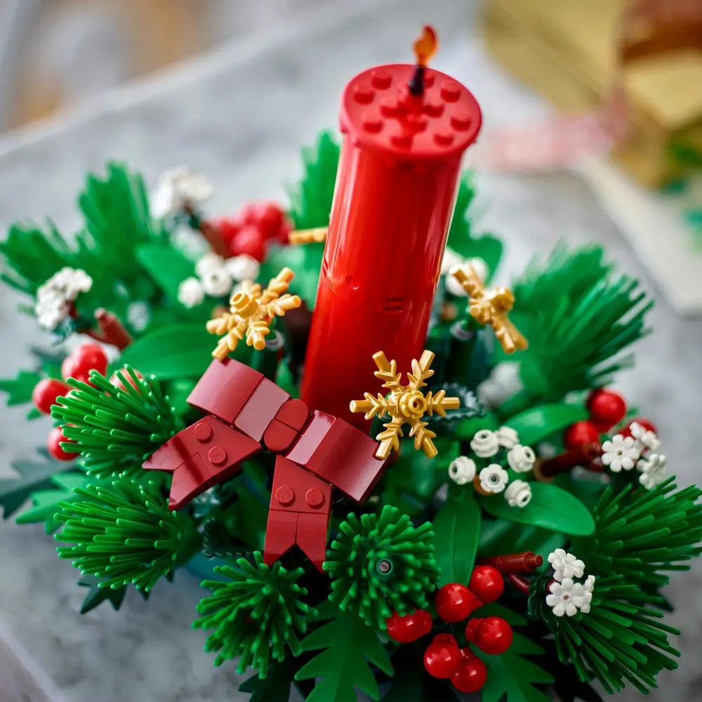 LEGO Christmas candle centerpiece featuring a tall red brick-built candle surrounded by green pine leaves, red berries, golden snowflakes, and a red bow for a festive holiday table display.