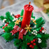 LEGO Christmas candle centerpiece featuring a tall red brick-built candle surrounded by green pine leaves, red berries, golden snowflakes, and a red bow for a festive holiday table display.