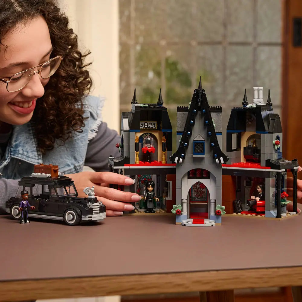 Builder assembling LEGO Wednesday Morticia’s Cottage set, placing detailed gothic furniture inside the open house, with Wednesday and Morticia minifigures nearby.