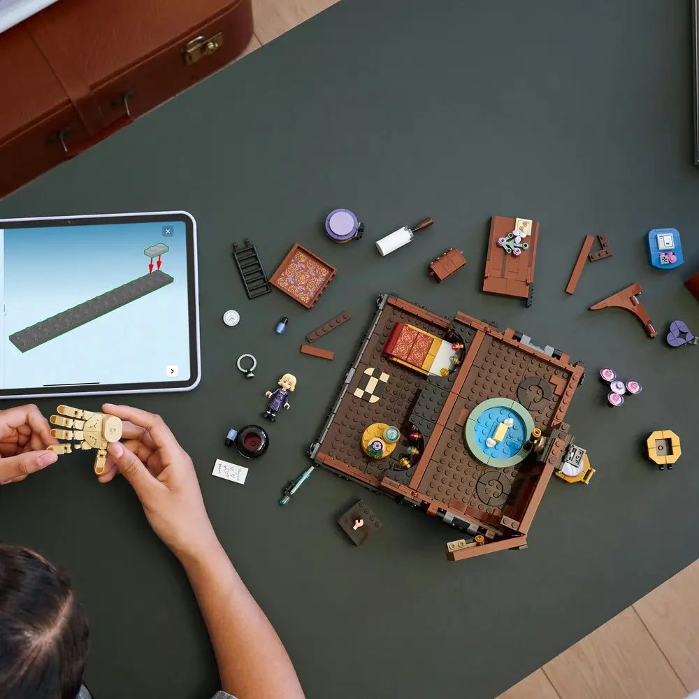 Overhead view of LEGO Wednesday Thing’s Apartment set being built, with brown and purple pieces, minifigures, accessories, and a tablet showing digital instructions beside the half-assembled model.