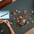 Overhead view of LEGO Wednesday Thing’s Apartment set being built, with brown and purple pieces, minifigures, accessories, and a tablet showing digital instructions beside the half-assembled model.