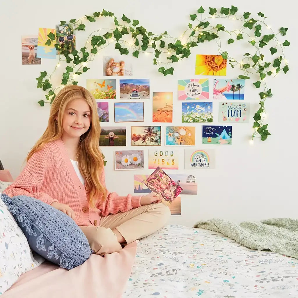 Smiling girl in a pink sweater sits on her bed with a photo collage and ivy fairy lights displayed on the wall behind her, creating a relaxing, creative bedroom atmosphere.
