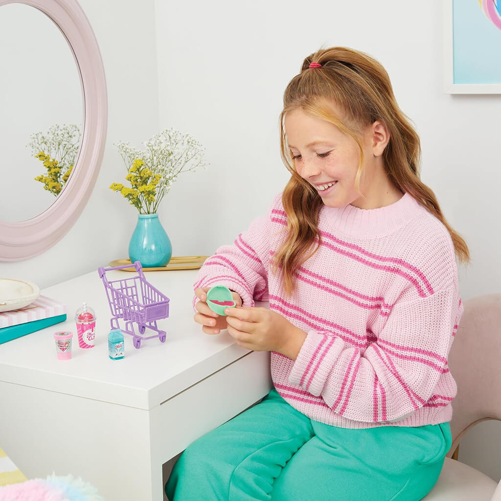 Smiling young girl wearing a pink striped sweater sitting at a white desk, playing with 3C4G Tiny Treats Trolley Lip Gloss Set items and miniature cart in a cozy bedroom.