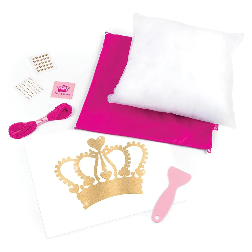 Juicy Couture DIY pillow kit components including a white pillow insert, hot pink cover, gold glitter crown transfer, pink embroidery thread, adhesive gems, and crafting spatula on a white background.