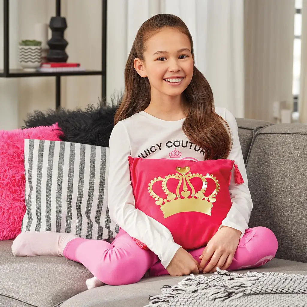 Smiling girl wearing a white Juicy Couture shirt and pink pants sits on a gray couch, holding a bright pink Juicy Couture pillow with a gold crown design surrounded by colorful pillows.