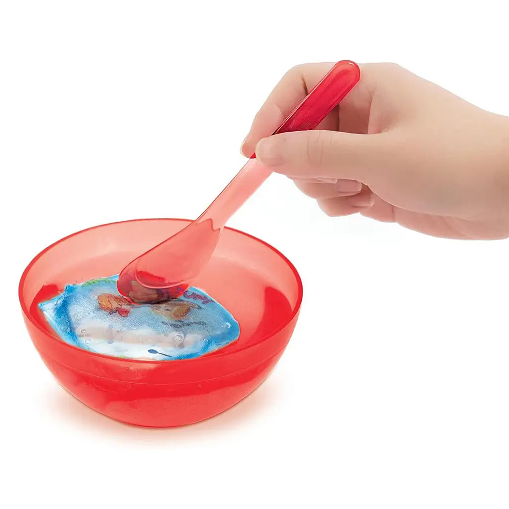 Hand holding a red spoon mixing a dissolvable blue pouch in a red bowl from the Kellogg’s Rice Krispies DIY Bracelet Kit, revealing colorful hidden beads as part of the craft process.