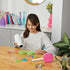 Smiling young girl using a hair dryer to shrink Make It Real Shrink Magic beads while crafting bracelets at a bright, colorful table setup.