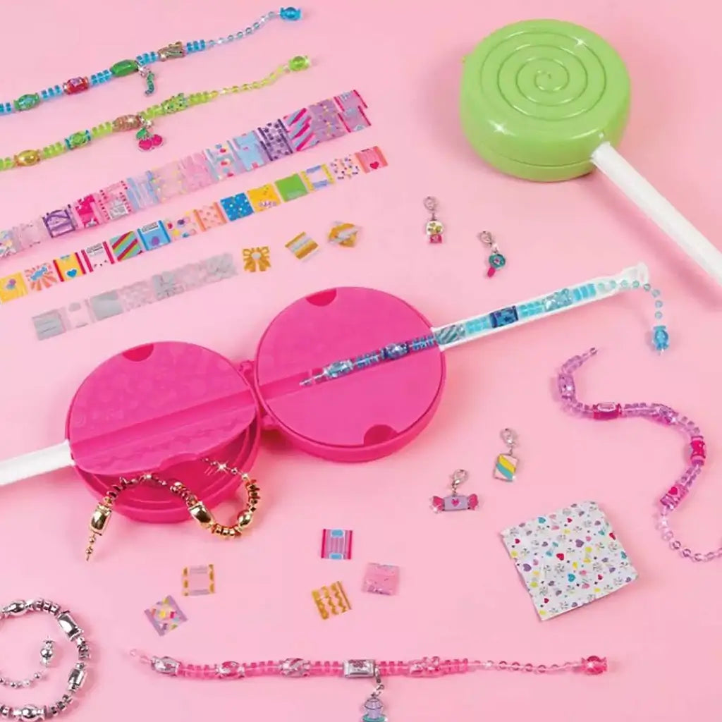 Flat lay of Make It Real Shrink Magic DIY Bracelet Kit on pink background with beads, wrappers, charms, and opened shrink lollipop tools arranged neatly.