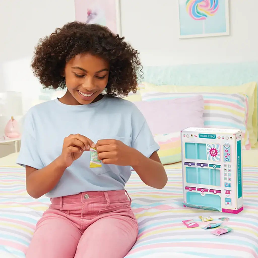 Smiling girl sitting on a striped bedspread opening a mini snack bag from the Make It Real Snackers Bracelet Kit vending machine, enjoying a creative playtime activity.