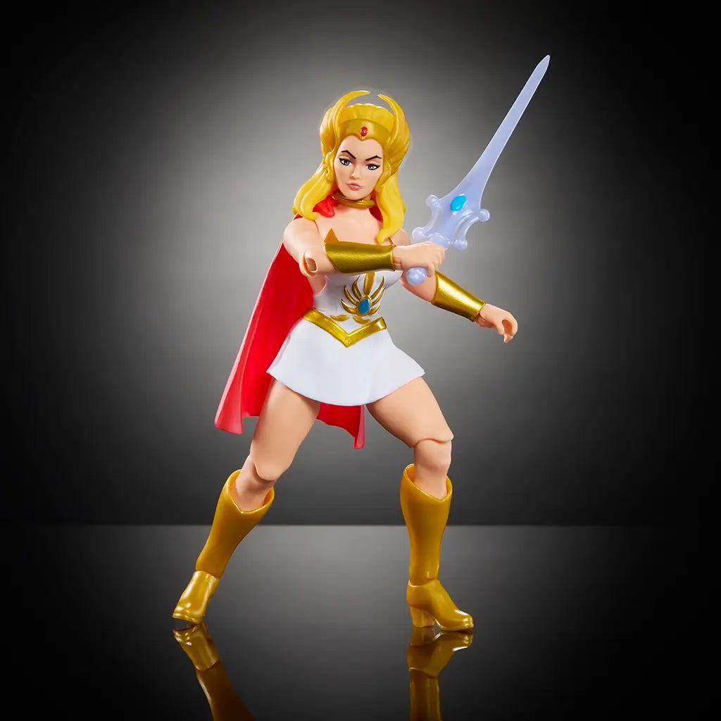 Masters of the Universe She-Ra action figure with long blonde hair, white and gold outfit, red cape, and silver sword, posed in dynamic battle stance on reflective black surface.
