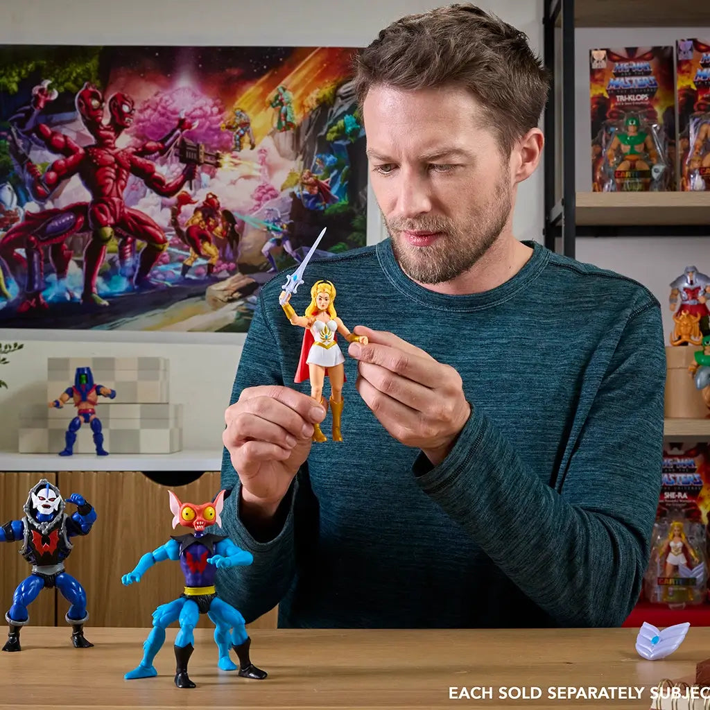 Person holding She-Ra action figure with sword and red cape, surrounded by other Masters of the Universe toys, in a colorful indoor display scene.