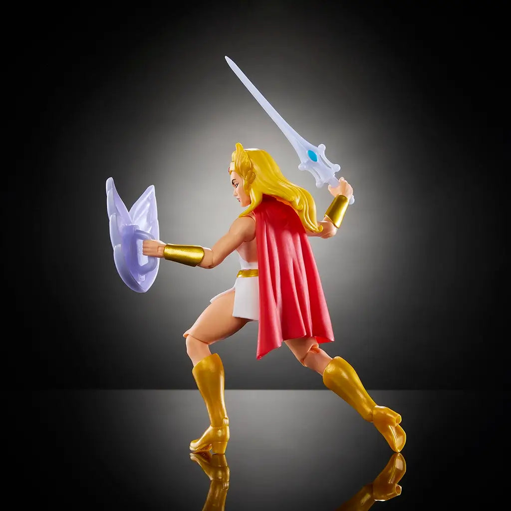 Side view of She-Ra Masters of the Universe figure with flowing blonde hair, white and gold attire, red cape, and sword raised for battle, holding white shield.