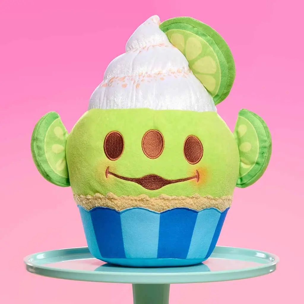 Disney Munchlings Pixar Toy Story Alien plush shaped like a lime cupcake with white whipped topping, lime slice ears, and a blue cupcake base, displayed on a pastel stand against a pink background.