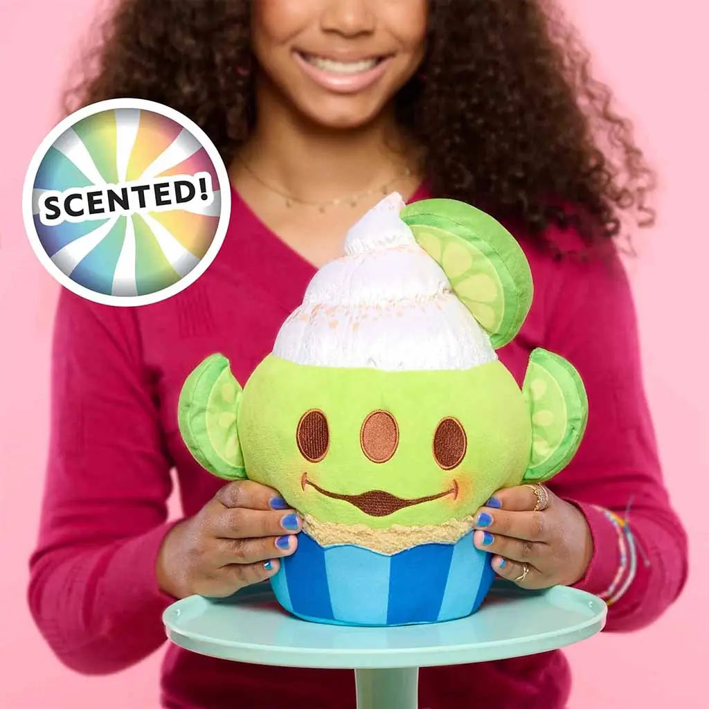 Smiling girl in red top holding a scented Disney Munchlings Alien plush designed as a green lime cupcake with brown eyes, white frosting, and lime slice ears, set against a bright pink background.