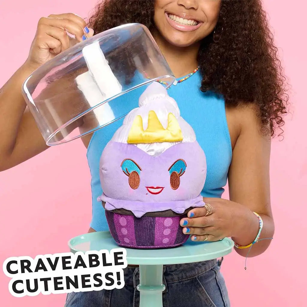 Smiling girl in blue tank top lifts a cake cover to reveal the Disney Munchlings Ursula plush shaped like a purple cupcake with a yellow crown and red smile, displayed on a mint cake stand with pink backdrop.