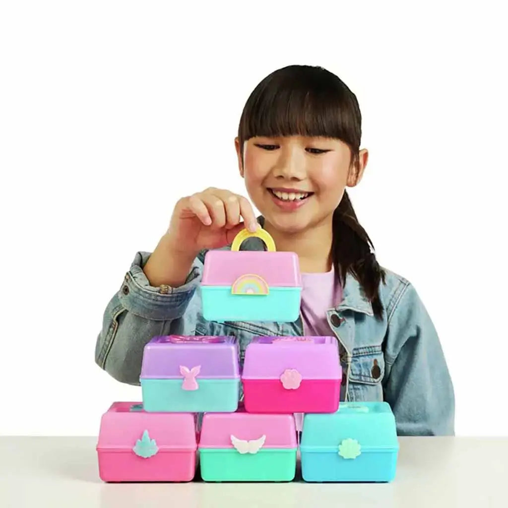 Smiling child stacking colorful Real Littles Micro Craft boxes in pink, purple, and teal with different charm handles, showing collection and playtime creativity on white background.
