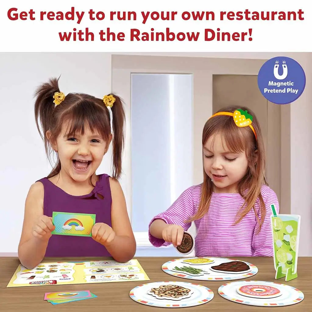 Two young girls smiling while playing with Skillmatics Rainbow Diner magnetic playset, arranging colorful pretend foods and play money on plates during imaginative restaurant playtime.