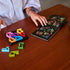 A person at a wooden table plays SmartGames IQ Square puzzle, arranging colorful block pieces while checking the illustrated challenge booklet for solutions.