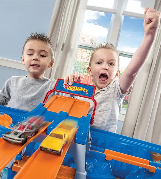 Two excited young boys playing with a Hot Wheels racetrack, launching toy cars down orange tracks. One boy raises his fist in excitement as the race unfolds.